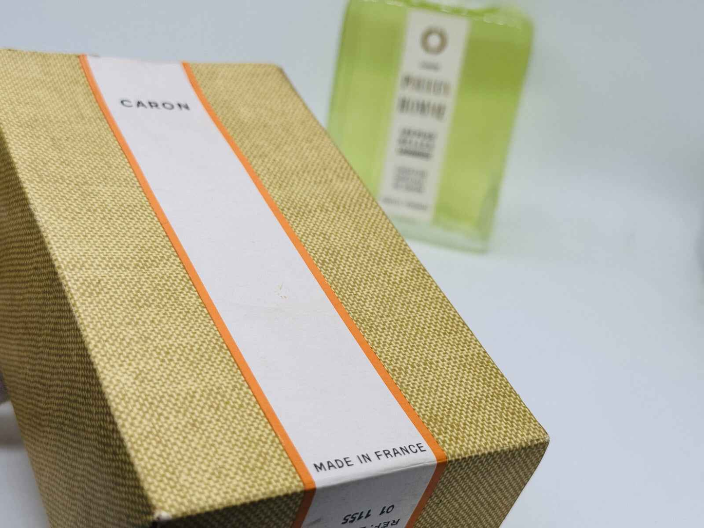 Scatola profumo Caron Pour Un Homme con etichetta bianca e arancione, sfondo sfocato di bottiglia verde